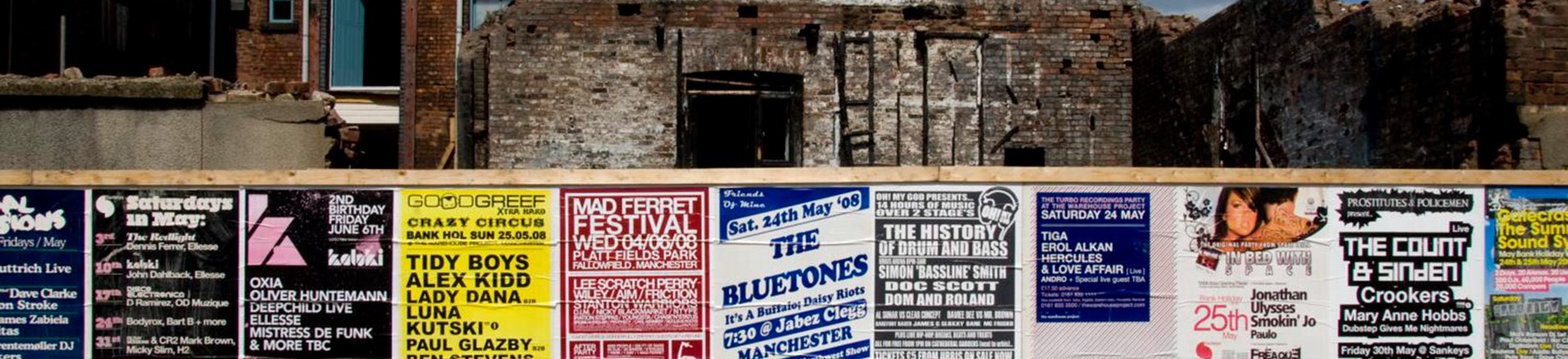 A wooden wall covered in colorful event posters with a partly demolished building and blue sky in the background.