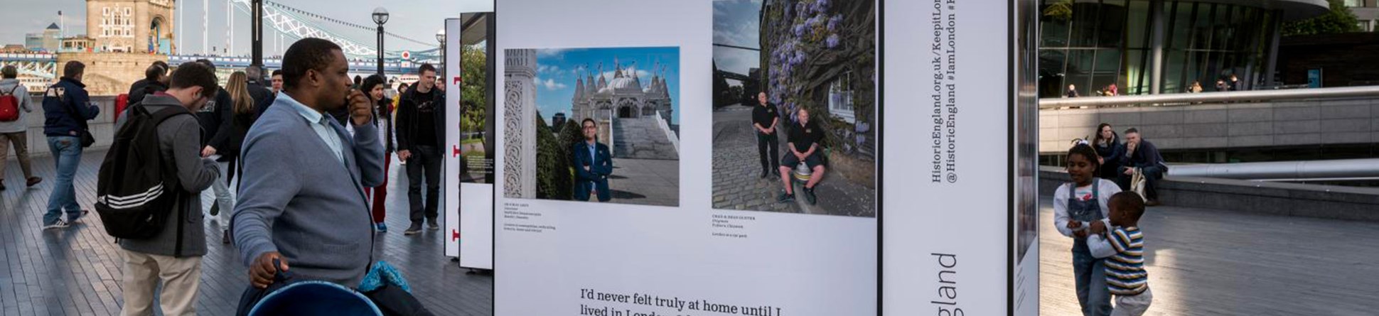 Outdoor exhibition near tower bridge, displaying panels with photos and texts, with people passing by and city hall in the background.