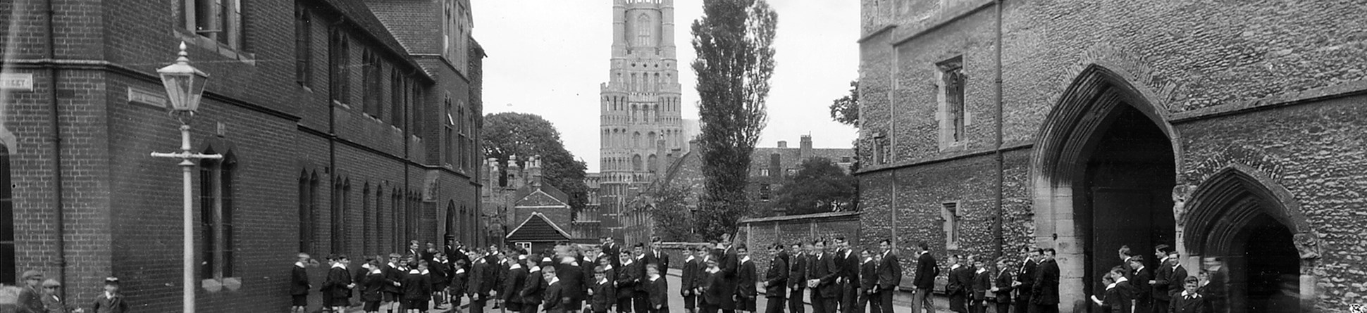 Archive black and white photograph of a group of schoolboys in the street between a brick building and a flint and stone medieval gatehouse, with a church tower in the background.