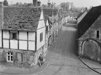 A black and white photograph of an elevated view of a woman walking down a street of stone and timber-framed houses.
