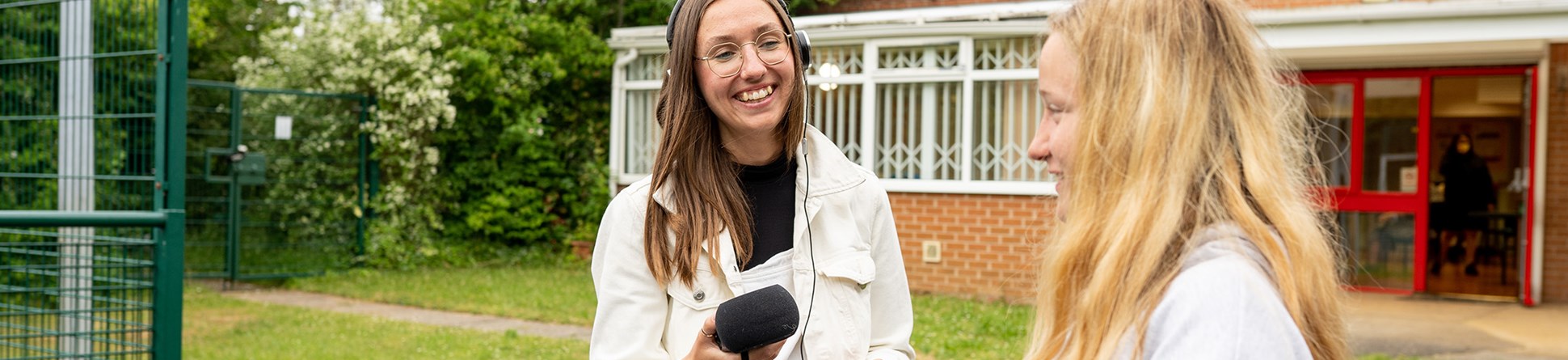 A person with long hair and round glasses dressed in white trousers and jacket holding a microphone and wearing headphones smiles at a person with long blonde hear wearing a grey sweatshirt outside a single storey brick building and grassy area.