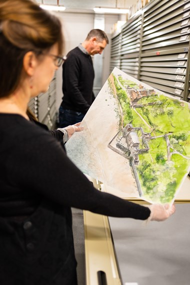 A colour photo of two people in an archive plans room. In the foreground, a person is holding up and looking at a colourful painting of a castle or fortified house.